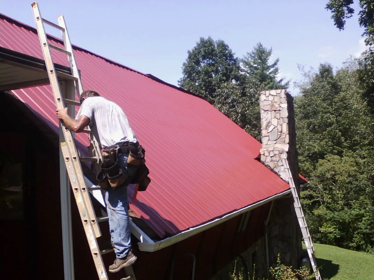 Roofer on a ladder beside a red metal roof and stone chimney — Soffit & Fascia Repair by Roofing Brothers in Carlton